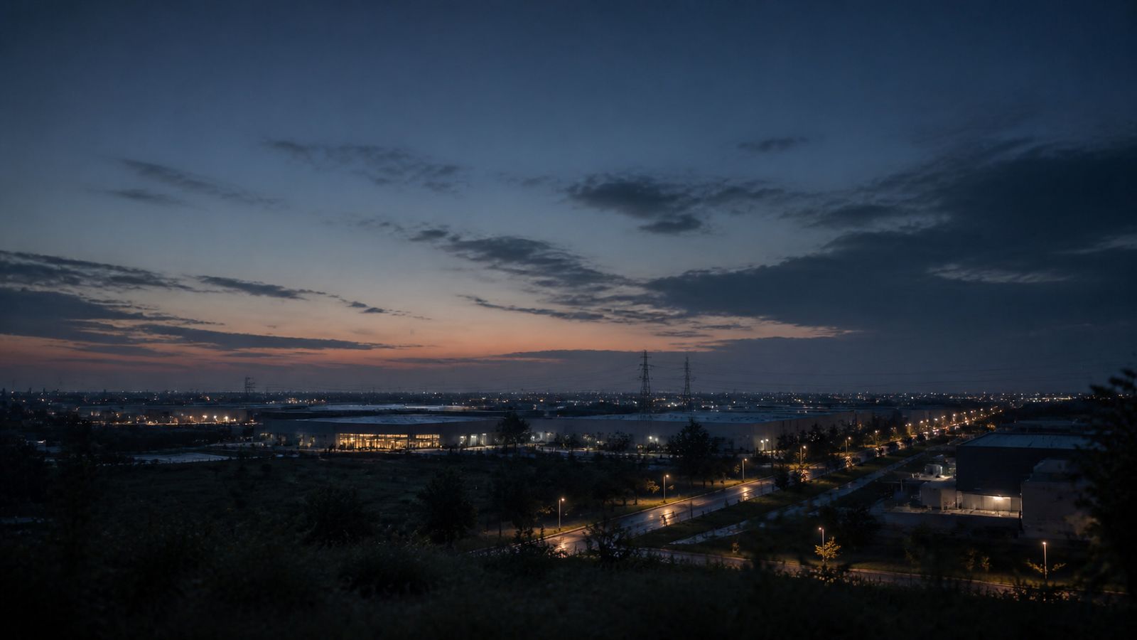 A wide blue-hour view across a low industrial landscape from slight elevation: a lit distribution building, lamp-lit roads, transmission pylons in the middle distance and a band of warm light along the horizon.