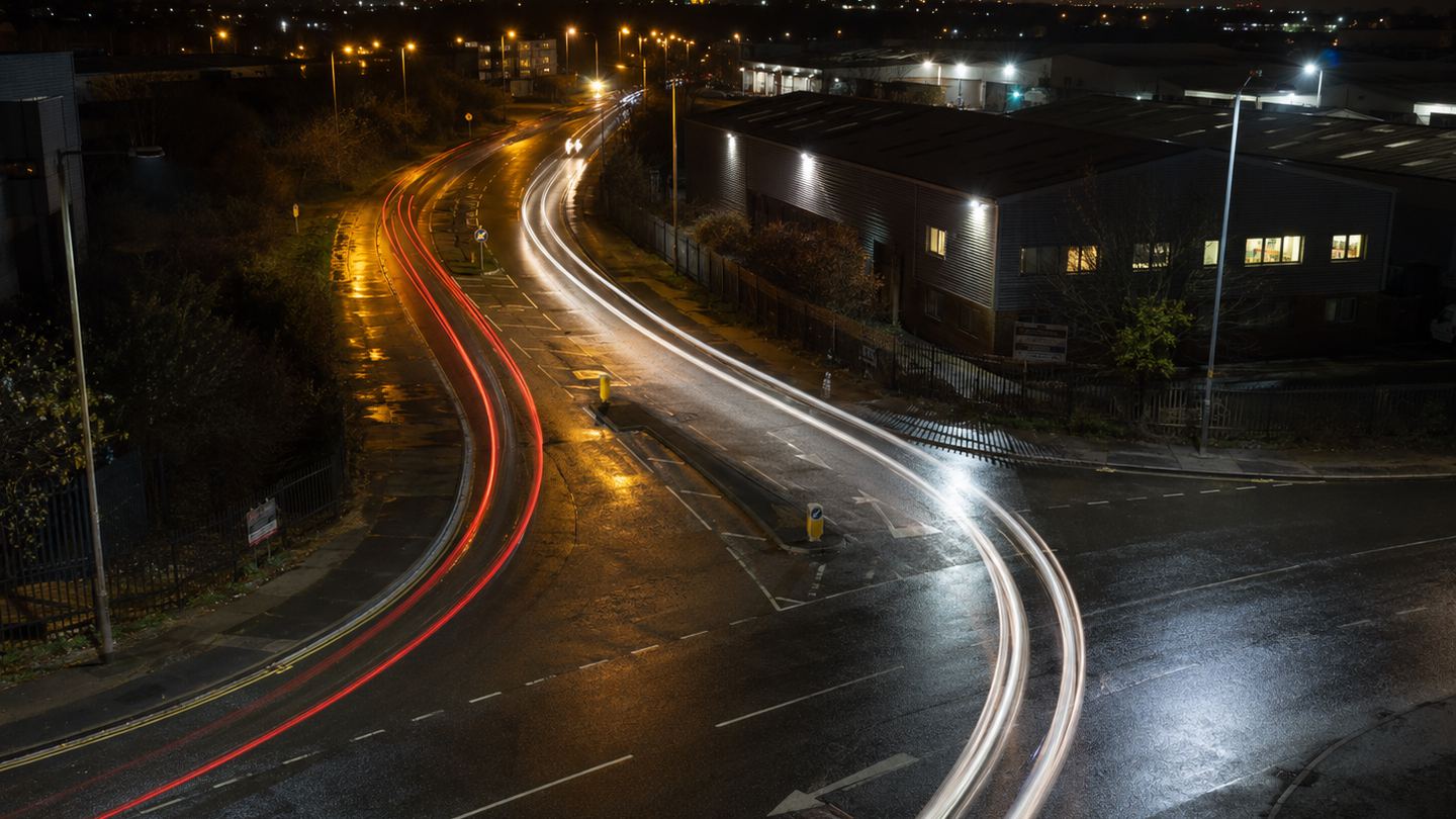 A long-exposure night view looking down on a wet road curving past a low industrial building, with red and white vehicle light trails drawn as continuous streaks through the bend and city lights along the horizon.